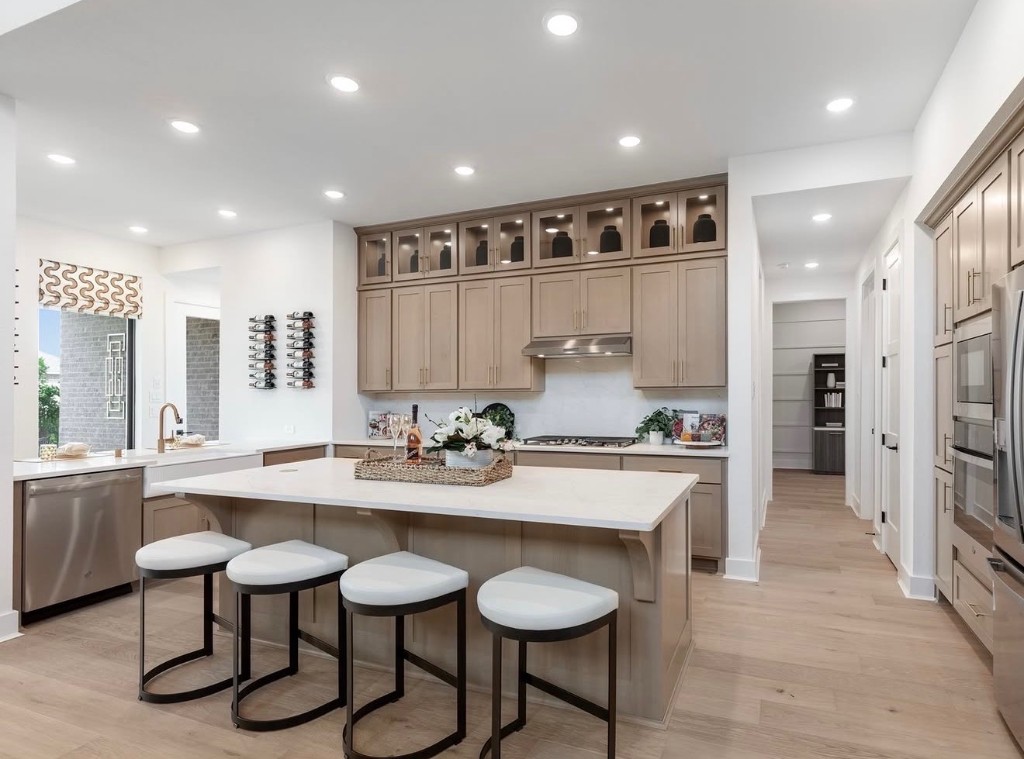 A bright modern kitchen with a large white island, beige cabinetry, barstools, and stainless steel appliances in an open, airy layout