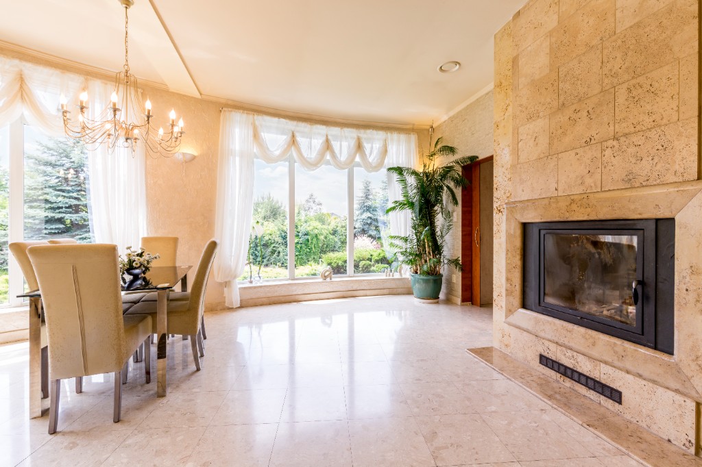 An elegant dining room with a stone fireplace, chandelier, and floor-to-ceiling windows overlooking a lush garden