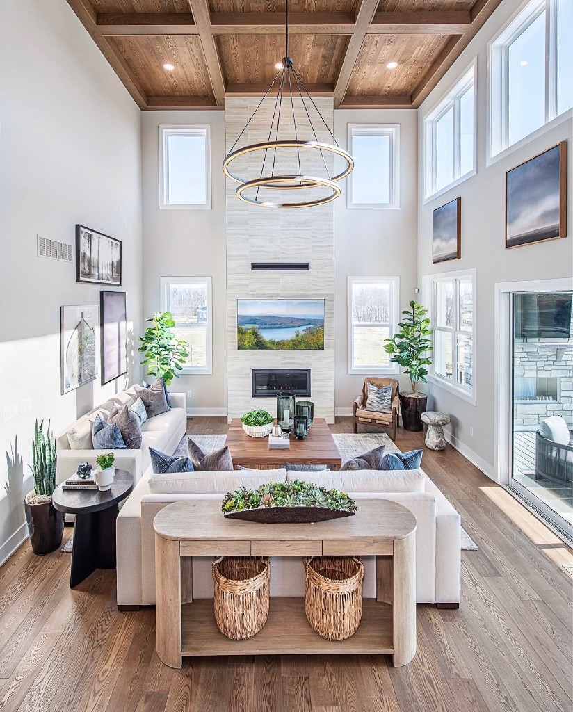 A bright luxury living room with a stone fireplace wall, coffered wood ceiling, halo chandelier, and floor-to-ceiling windows filling the space with natural light