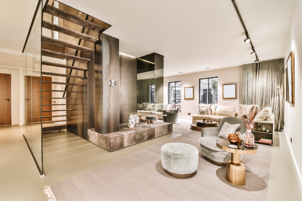 A luxurious modern home interior featuring a glass-railed staircase, a plush cream sofa, gray velvet armchairs, and elegant gold accents in a bright open-concept living room