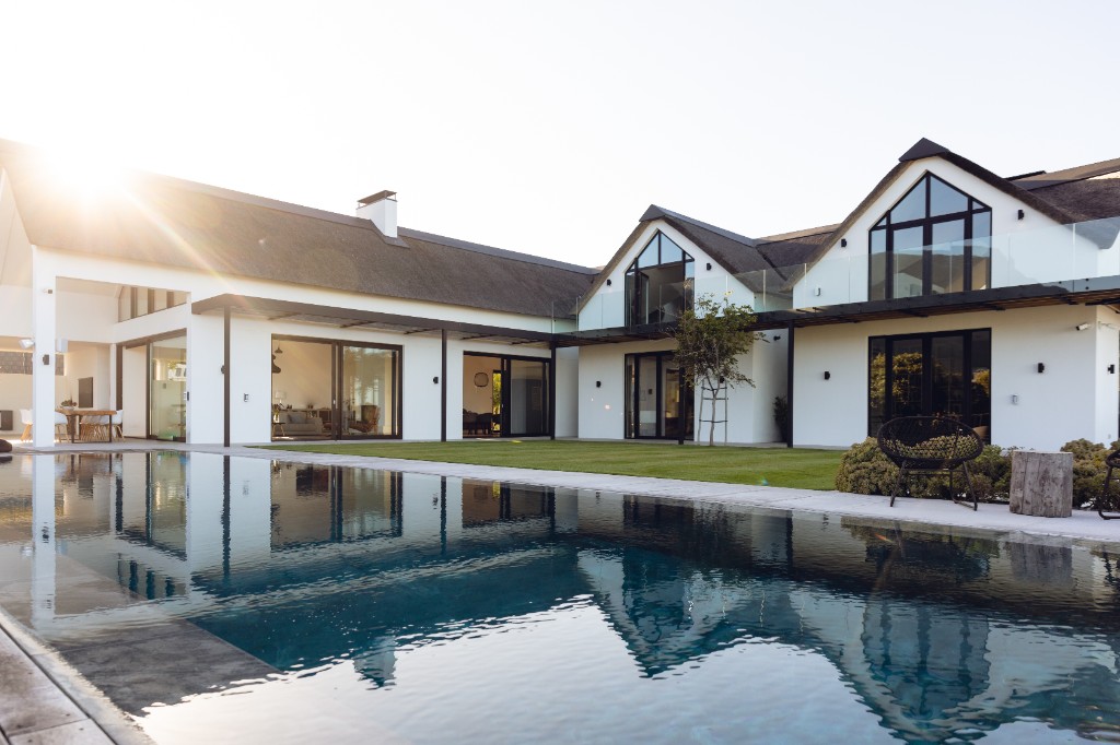 A luxurious modern white house with a dark gabled roof and large glass windows, featuring a swimming pool reflecting the architecture at golden hour